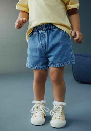 Lille barn iført gul skjorte, blå denimshorts, hvide sokker og cremefarvede høje sneakers, stående på en grå gulv.