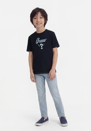 Chłopiec stoi i się uśmiecha, ubrany w czarną koszulkę Guess, jasnoniebieskie dżinsy oraz granatowe buty wsuwane, na jednolitym białym tle.