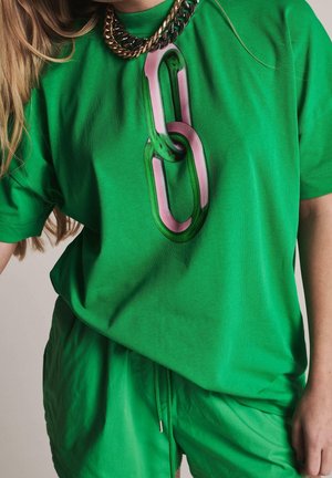 Kobieta ubrana w zielony strój z dużym nadrukiem łańcucha na koszulce i masywnym naszyjnikiem łańcuchowym, z długimi blond włosami widocznymi.