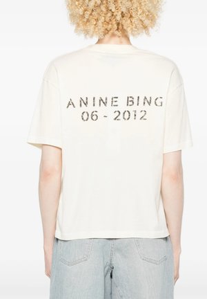 Persona con capelli biondi ricci indossa una maglietta bianca con "ANINE BING 06-2012" stampato sulla schiena e pantaloni di jeans azzurri chiari.