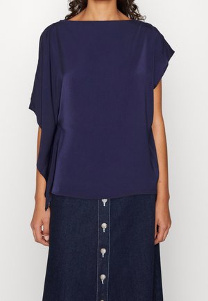 Kvinde iført en løs mørkeblå kortærmet bluse med bådudskæring og et mørkt knapforan denimskørt.