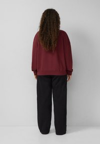 Een persoon met lang krullend haar staat tegenover een witte muur, gekleed in een donkerrode trui en losse zwarte broek.