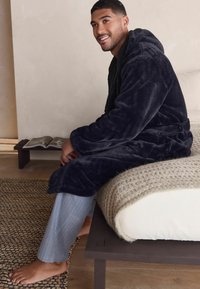 Navy plush robe with a hood, belted at the waist, worn over light gray pinstriped pants. Textured beige blanket on a bed.