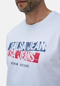 Weißes T-Shirt mit kurzen Ärmeln, das mit einem blauen und roten grafischen Textdruck versehen ist, der "SALSA JEANS" und "DENIM ARTISANS" in einer fetten Schriftart zeigt.