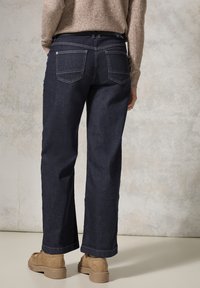 Vaqueros de mezclilla azul oscuro con corte holgado, que cuentan con bolsillos traseros con costuras visibles, combinados con calzado marrón.