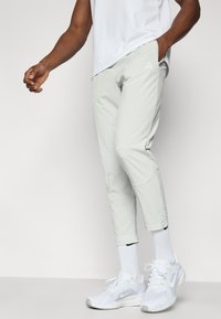 Αθλητικό παντελόνι ανοιχτό γκρι με στενότερα μπατζάκια, με λογότυπο στον μηρό, συνδυασμένο με λευκά αθλητικά παπούτσια και ριμπ κάλτσες αστραγάλου.