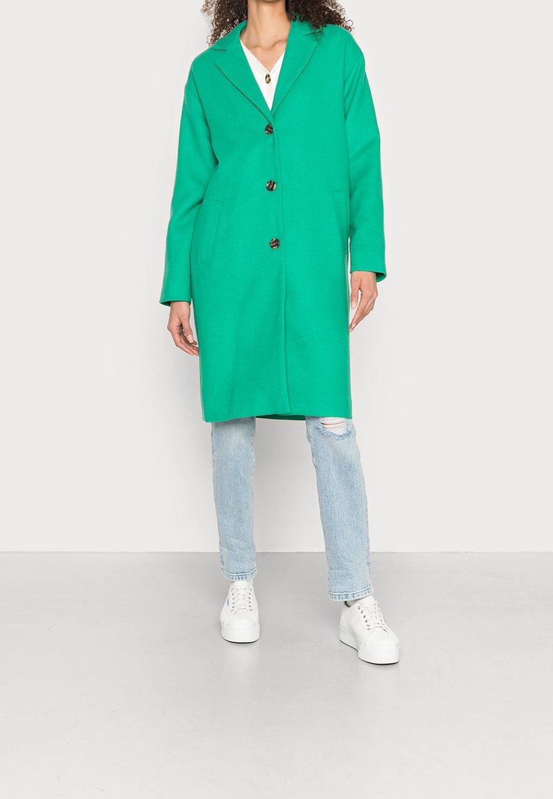 Grøn uldblandet frakke med skrå krave, fire forreste knapper og to sidelommer, båret over lyseblå slidte jeans og hvide sneakers.