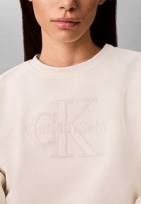 Kvinne iført en kremfarget Calvin Klein-genser med hevet teksturert logo midt på brystet, nøytral bakgrunn synlig.