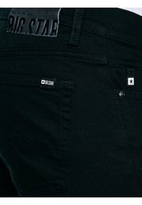 Fekete farmernadrág hátuljának zsebének közeli képe, amelyen látható a „BIG STAR” márkajelzés és a varrás részletei.