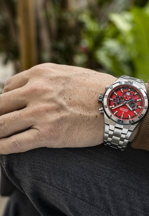 Montre en acier inoxydable avec un cadran rouge, des sous-cadrans noirs et des marqueurs lumineux. Équipée d'un bracelet en métal et de trois boutons de chronographe.