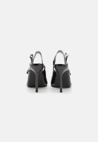 Een paar zwarte lakleren stilettohakken gezien vanaf de achterkant, met slingback-bandjes met witte Karl Lagerfeld-branding.