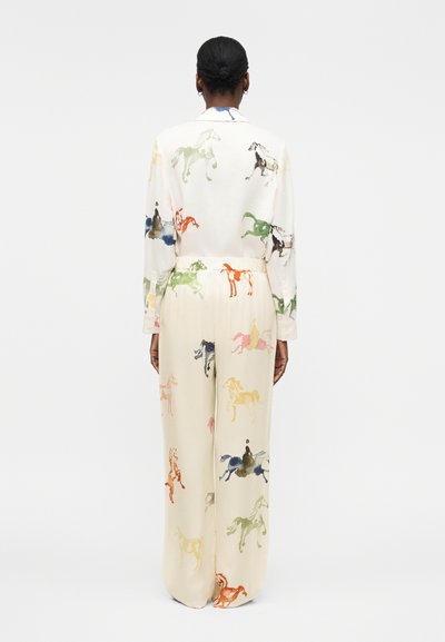 Γυναίκα στέκεται με την πλάτη γυρισμένη, φοράει κοστούμι σε κρεμ χρώμα με πολύχρωμες εκτυπώσεις αλόγων και αναβατών σε σακάκι και παντελόνι.