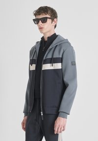 Giacca con cappuccio grigia e blu navy, dotata di chiusura con zip, strisce a contrasto e una toppa con logo. Indossata sopra un top nero a collo alto.