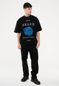 Black oversized t-shirt with a blue graphic design featuring the text "NEW ORDER SUBSTANCE 1987". Paired with black trousers and boots.
