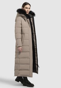 Hosszú bézs puffertakaró kapucnival, fekete szőrmével díszítve, tűzött mintázattal, dupla oldalsó zsebekkel és karcsú siluettal, fekete ruházat felett viselve.