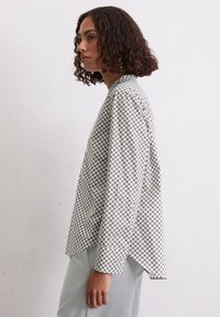 Chemise à manches longues blanche avec un motif géométrique marron, présentant une coupe décontractée et un ourlet légèrement arrondi. Tissu doux et coupe ample.