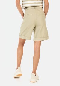 Khakifarbene Leinen-Shorts mit umgeschlagenen Säumen, mit Gesäßtaschen und einer lockeren Passform. Getragen mit weißen Sneakers vor einem neutralen Hintergrund.