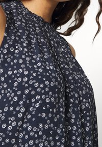 Marinblus utan ärmar med vit blommönster, plisserad design och rund halsringning med en subtil volangdetalj. Mjuk textur.