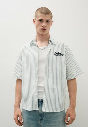 Junger Mann mit kurzen Haaren, der ein weißes Kurzarmhemd mit Nadelstreifen über einem weißen Tanktop und blauen Jeans vor schlichtem Hintergrund trägt.