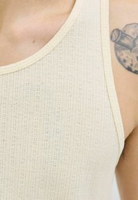Canotta a coste color crema con scollatura rotonda, caratterizzata da sottili righe orizzontali e una superficie texture. Un tatuaggio è parzialmente visibile.