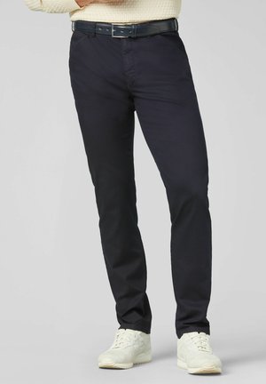 Mann trägt schwarze Slim-Fit-Hose, schwarzen Ledergürtel, weiße Sneakers und cremefarbenen Strickpullover, steht vor einem schlichten Hintergrund.