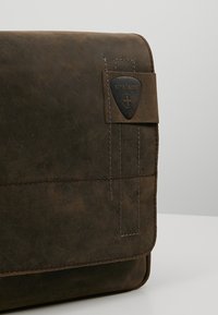 Borsa a tracolla in pelle marrone con chiusura a pattina, cuciture a contrasto e un'etichetta con logo nera. Texture liscia con finitura opaca.