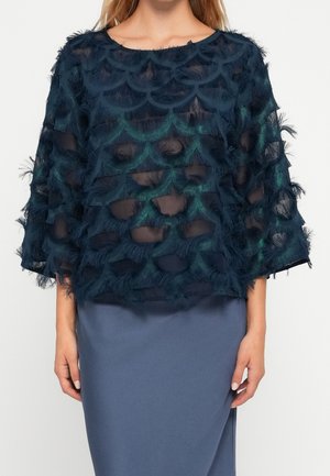 Mujer que lleva un top azul marino transparente con patrones texturizados semejantes a escamas sobre una capa base color azul verdoso y una falda azul sólida.