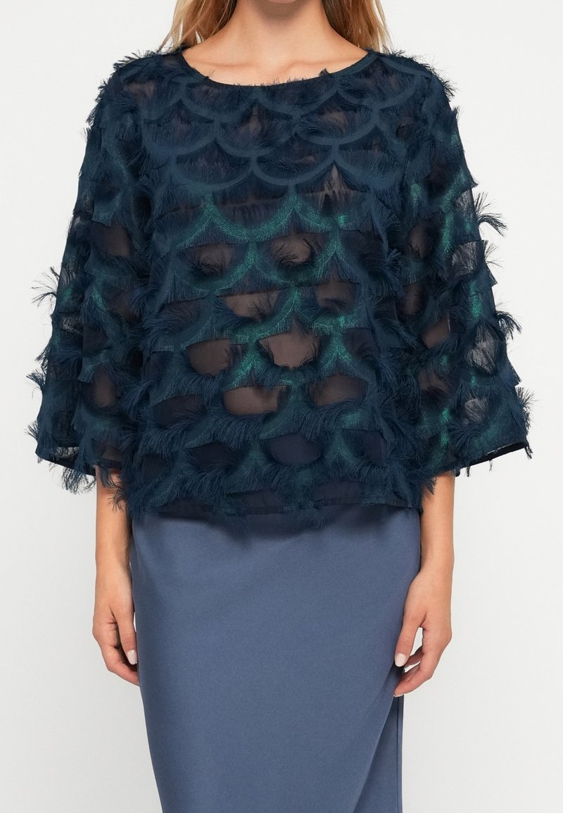 Femme portant un haut transparent bleu marine avec des motifs texturés en forme d'écailles sur un sous-vêtement bleu sarcelle et une jupe bleue unie.