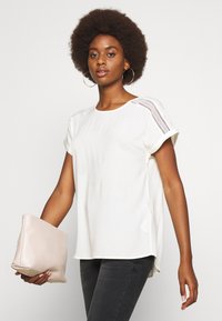 Top branco de manga curta com decote redondo, apresentando detalhes em renda nos ombros. Combinado com jeans justos e uma carteira rosa clara.