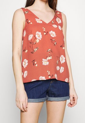 Vrouw draagt een mouwloos, roestkleurig bloesje met witte en roze bloemen en donkerblauwe opgerolde spijkershorts.