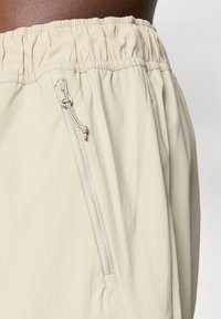 Shorts légers beiges avec une taille élastique, dotés d'une poche latérale zippée et d'une texture lisse, légèrement froissée.