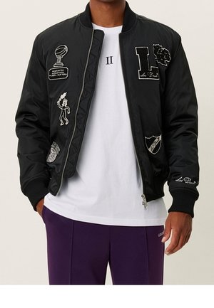 Mann trägt schwarze Bomberjacke mit weißen Aufnähern, weißes T-Shirt mit dem Text "II" und lila Hose, steht mit teilweise sichtbaren Händen.