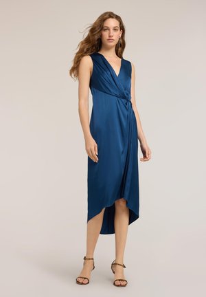 Femme portant une robe portefeuille bleu roi sans manches avec un ourlet asymétrique et des sandales marron à brides, debout devant un fond uni.