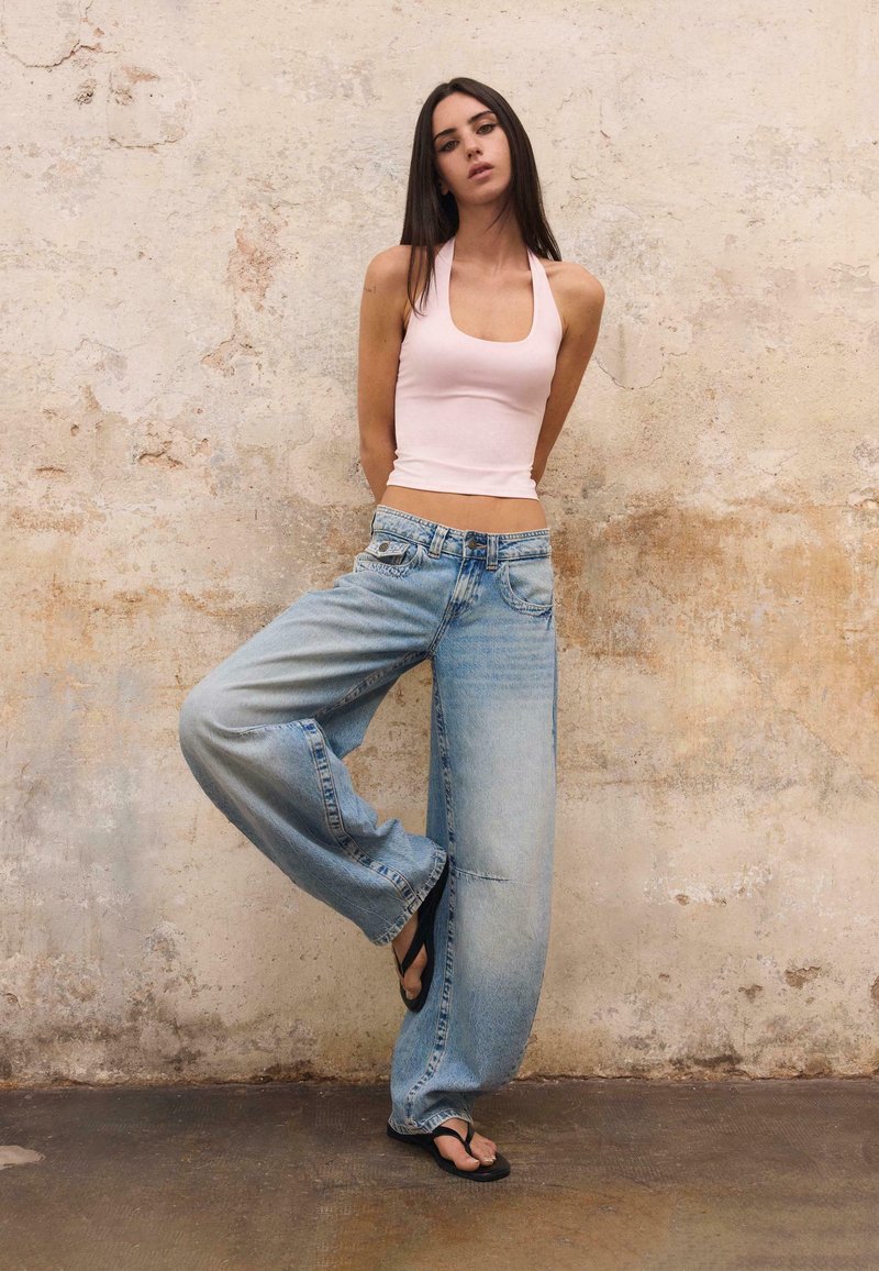 Femme portant un haut dos nu rose clair, un jean bleu clair à jambes larges et des tongs noires, debout avec une jambe levée contre un mur texturé.