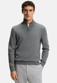 Pull-over gris avec col à fermeture éclair quart, poignets côtelés, conçu dans un tissu en maille douce, associé à un pantalon gris clair.