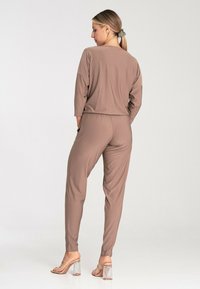 Brown, lightweight two-piece loungewear set featuring a relaxed long-sleeve top and tapered pants with an elastic waistband, paired with clear heels.
