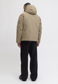 Beige wasserdichte Kapuzenjacke mit geradem Schnitt, kombiniert mit lockeren schwarzen Hosen. Minimalistisches Design, glatte Stoffstruktur und keine sichtbaren Beschläge.