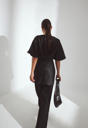 Čierny saténový top s prehnanými rukávmi, kombinovaný s voľnými čiernymi nohavicami. Postava drží čiernu tašku, je videná zozadu na svetlom pozadí.