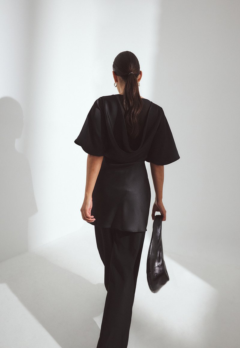 Čierny saténový top s prehnanými rukávmi, kombinovaný s voľnými čiernymi nohavicami. Postava drží čiernu tašku, je videná zozadu na svetlom pozadí.