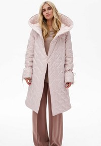 Jasnoróżowy pikowany płaszcz z długim rękawem i oversize'owym kapturem, z elastycznymi mankietami i zapięciem na napy. W połączeniu z beżowymi spodniami.