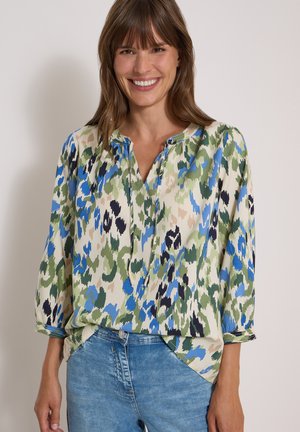 Femme souriante portant une blouse ample avec un imprimé abstrait vert, bleu, noir et beige ainsi qu'un jean en denim bleu clair.