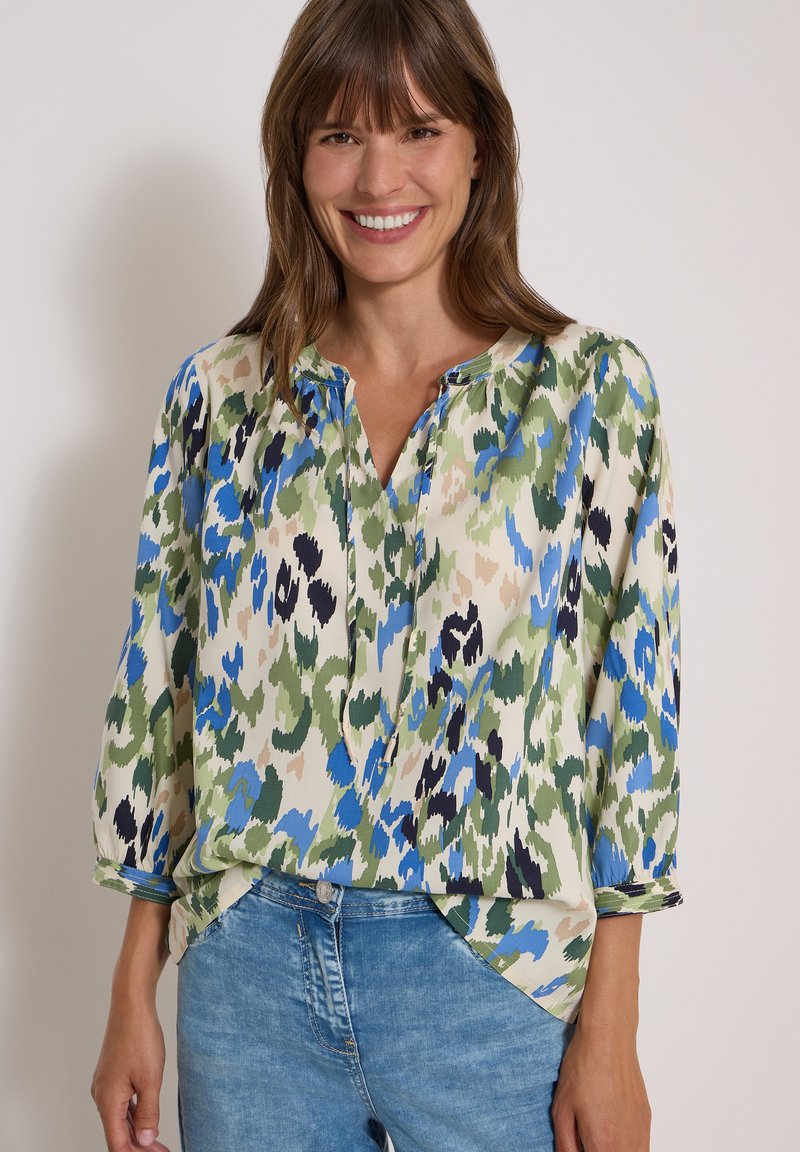 Femme souriante portant une blouse ample avec un imprimé abstrait vert, bleu, noir et beige ainsi qu'un jean en denim bleu clair.
