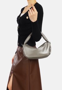 Borsa a spalla grigio metallizzato con design curvo, texture liscia e manico singolo. Indossata con un cardigan nero a coste e una gonna marrone.