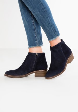 Person iført mørkeblå ruskind ankle støvler med sidelommer og lave træblokhæle, kombineret med blå jeans med frynset kant.