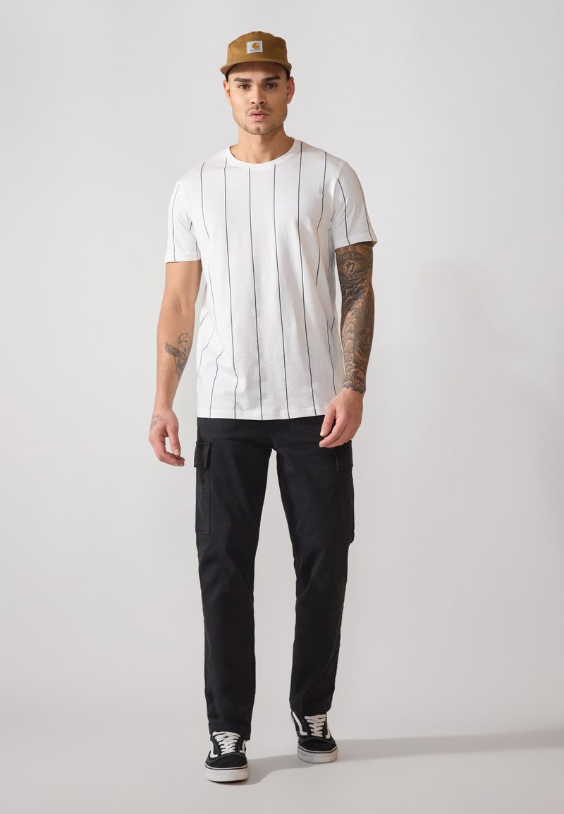 Camiseta blanca de manga corta con rayas verticales negras, combinada con pantalones cargo negros y zapatillas en blanco y negro. Gorra marrón en la cabeza.
