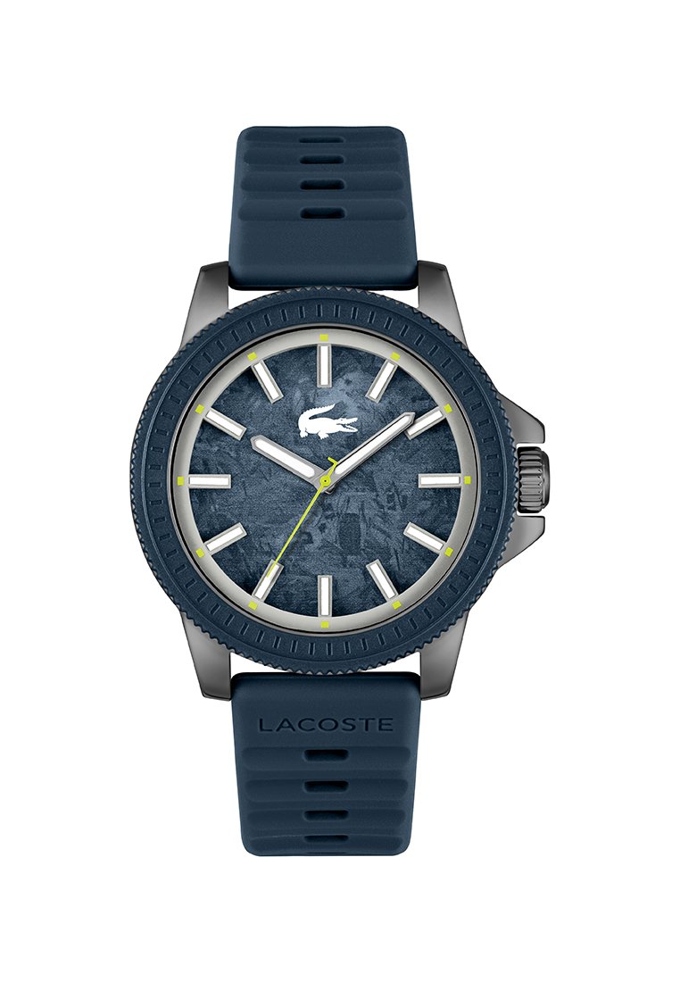 Montre Lacoste avec un bracelet en caoutchouc bleu, cadran texturé bleu foncé et index des heures argentés. Présente une aiguille des secondes jaune et un détail de logo.