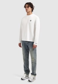 Langärmliges weißes Hemd mit einem kleinen schwarzen Logo auf der Brust, kombiniert mit hellblauen, verwaschenen Jeans und silbernen Sportschuhen.