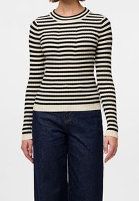 Femme portant un pull à manches longues rayé noir et blanc et un jean taille haute bleu foncé, debout devant un fond blanc uni.