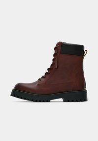 Bota de lace-up em couro castanho com colarinho acolchoado preto, sola grossa preta e puxador amarelo, mostrada de lado sobre fundo branco.