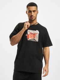 Sort bomulds T-shirt med et grafisk motiv af et revet papirmotiv med orange og hvide accenter. Korte ærmer og løst fit, som viser syningsdetaljer.